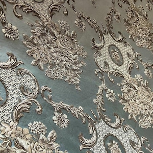 Può includere: Primo piano di un tessuto blu chiaro con un motivo damascato argento e oro. Il motivo presenta intricati disegni floreali e volute.