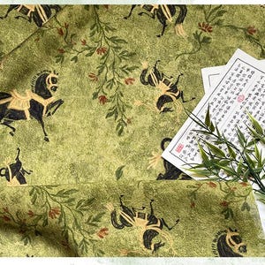 Może przedstawiać: Zbliżenie zielonej tkaniny z powtarzającym się wzorem czarnych koni i element&oacute;w kwiatowych. Tkanina ma teksturowany wygląd. Kawałek papieru z azjatyckimi znakami i gałązkami bambusa znajduje się w rogu.