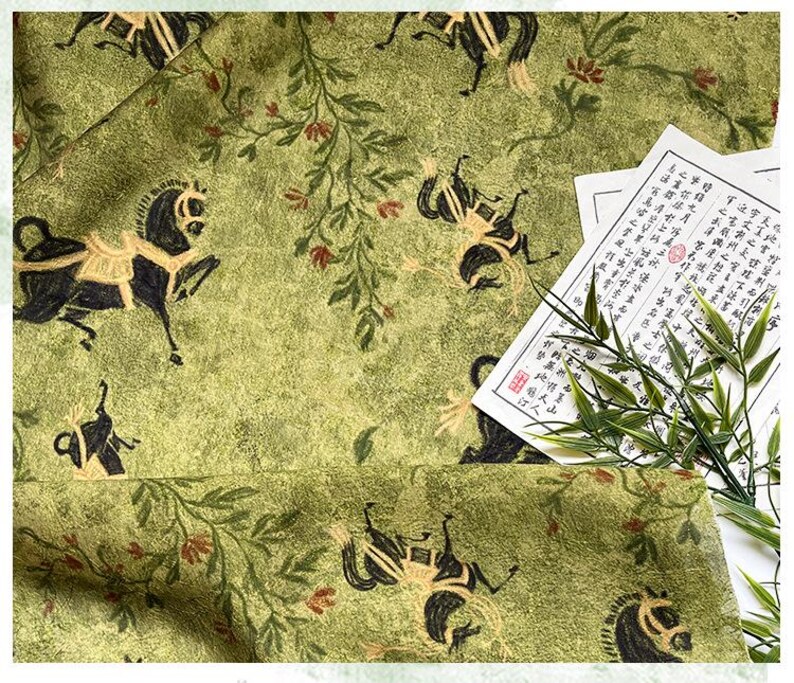 Może przedstawiać: Zbliżenie zielonej tkaniny z powtarzającym się wzorem czarnych koni i element&oacute;w kwiatowych. Tkanina ma teksturowany wygląd. Kawałek papieru z azjatyckimi znakami i gałązkami bambusa znajduje się w rogu.