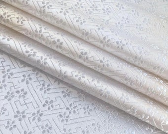 Op de afbeelding: Witte stof met een subtiel geometrisch patroon en bloemenontwerp. De stof heeft een gladde, zijdeachtige textuur.