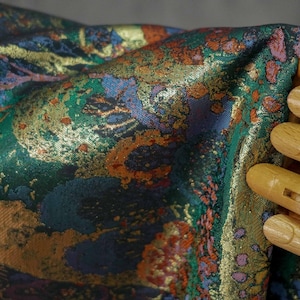 Puede incluir: Primer plano de una tela con un patr&oacute;n complejo. La tela presenta una mezcla de colores, incluyendo verde, azul, naranja y dorado. El dise&ntilde;o parece ser floral o abstracto. La tela est&aacute; siendo sostenida por una mano de madera.