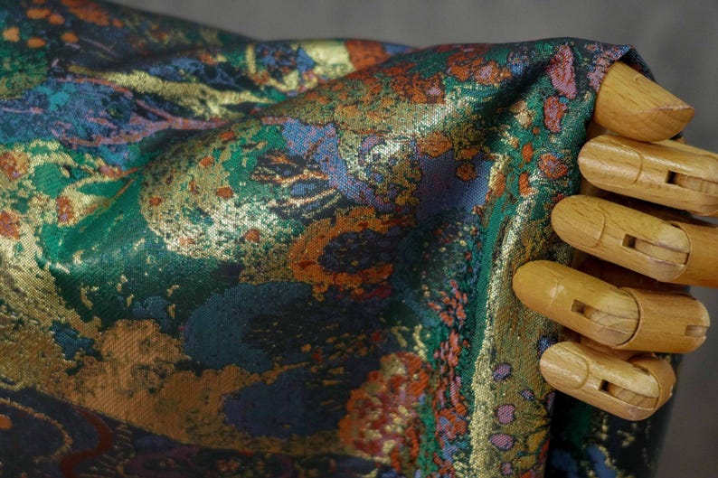 Puede incluir: Primer plano de una tela con un patr&oacute;n complejo. La tela presenta una mezcla de colores, incluyendo verde, azul, naranja y dorado. El dise&ntilde;o parece ser floral o abstracto. La tela est&aacute; siendo sostenida por una mano de madera.
