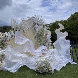 Könnte beinhalten: Eine Hochzeitszeremonie-Kulisse im Freien mit weißen Blumen, fließendem weißen Stoff und einer cremefarbenen Struktur. Die Szene spielt sich auf einer Rasenfläche mit Bäumen und einem bewölkten Himmel im Hintergrund ab. Eine schwarze Leiter ist sichtbar.