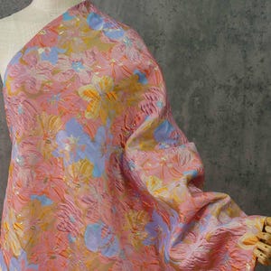 Op de afbeelding: Een bloemig stofstaal in roze, oranje, geel en blauw. De stof heeft een textuur en een herhalend patroon van gestileerde bloemen. De stof is over een mannequin gedrapeerd, wat de drape en textuur laat zien.