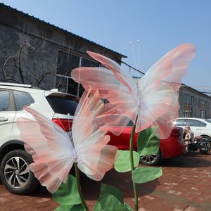 May include: Two large, decorative butterfly sculptures with pink and white translucent wings. The butterflies are mounted on green stems with large green leaves. The background includes cars and a building under a blue sky.