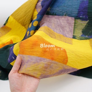Può includere: Un tessuto giallo con una stampa astratta colorata. Il tessuto ha una superficie testurizzata ed è piegato a metà. Il testo "Bloom 设计师面料" è stampato sul tessuto.