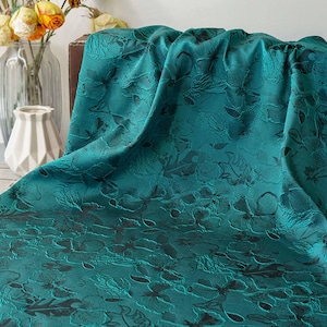 May include: Teal fabric with a floral pattern. The fabric is draped over a surface, showcasing its texture and design. The floral pattern is darker than the teal background, creating a contrast. The fabric is likely used for home decor or fashion.