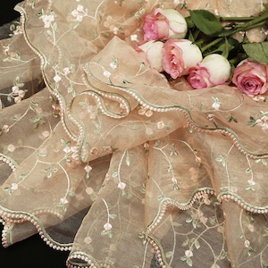 May include: A sheer peach-colored fabric with delicate floral embroidery and a scalloped edge. The fabric is draped over a bouquet of pink roses.