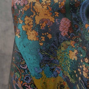 Puede incluir: Primer plano de una tela con un patr&oacute;n floral detallado. El dise&ntilde;o presenta una mezcla de colores, incluyendo verde azulado, dorado, morado y verde. La tela tiene una apariencia texturizada, con un dise&ntilde;o tipo paisley en la esquina inferior izquierda.