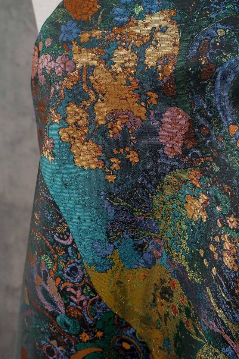 Puede incluir: Primer plano de una tela con un patr&oacute;n floral detallado. El dise&ntilde;o presenta una mezcla de colores, incluyendo verde azulado, dorado, morado y verde. La tela tiene una apariencia texturizada, con un dise&ntilde;o tipo paisley en la esquina inferior izquierda.
