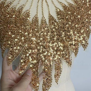 Könnte beinhalten: Nahaufnahme einer Applikation aus goldenen Pailletten und Perlen. Das Design zeigt ein fächerartiges Muster mit aufwendigen Details. Die Pailletten sind dicht gepackt und erzeugen einen schimmernden Effekt. Die Applikation befindet sich auf einem durchsichtigen, cremefarbenen Stoff.