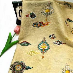 Op de afbeelding: Beige stof met een getextureerd oppervlak en een herhalend patroon van kleurrijke wolken- en bloemmotieven. Het ontwerp bevat turquoise, gele, rode en groene accenten. De stof is gevouwen en toont de herhaling van het patroon.