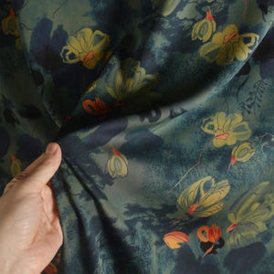 Op de afbeelding: Stof met een bloemmotief in groen, blauw, geel en oranje. Het ontwerp toont gestileerde bloemen en bladeren. De stof heeft een zachte, vloeiende uitstraling, geschikt voor kleding of woondecoratieprojecten.