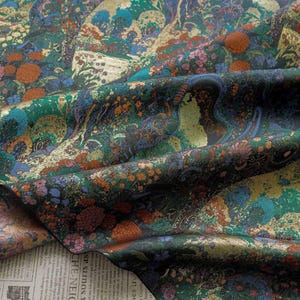 Puede incluir: Una tela detallada con un dise&ntilde;o de paisaje. La tela presenta una mezcla de colores, incluyendo verde azulado, dorado, naranja y morado. El dise&ntilde;o incluye elementos florales y arb&oacute;reos, creando una apariencia rica y texturizada. La tela es probablemente seda o un material similar.