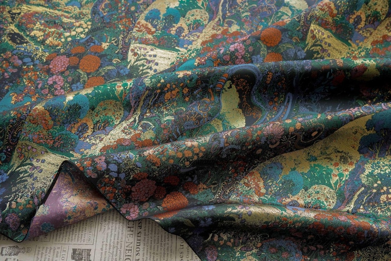 Puede incluir: Una tela detallada con un dise&ntilde;o de paisaje. La tela presenta una mezcla de colores, incluyendo verde azulado, dorado, naranja y morado. El dise&ntilde;o incluye elementos florales y arb&oacute;reos, creando una apariencia rica y texturizada. La tela es probablemente seda o un material similar.