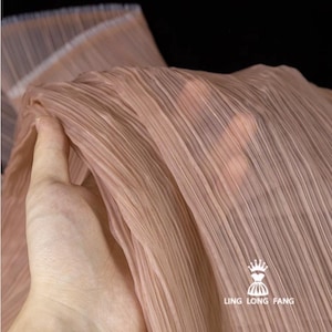 May include: Close-up of a pleated fabric in a soft, muted peach color. The fabric has a delicate, sheer quality with vertical folds. The hand is holding the fabric. The text "LING LONG FANG" is visible in the lower right corner.