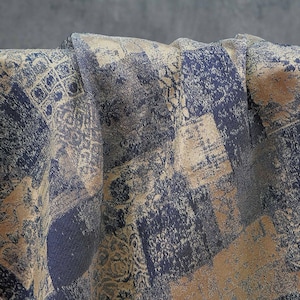 Pode incluir: Close-up de um tecido dobrado com um design de patchwork. O tecido apresenta uma mistura de quadrados azul marinho, bege e cinza com padrões texturizados. A estética geral é vintage e artística, adequada para decoração de casa ou moda.