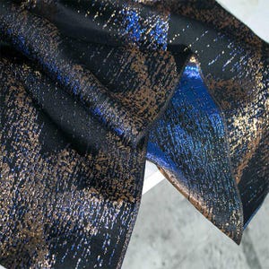 Op de afbeelding: Close-up van een zwarte stof met een metallic blauw en gouden patroon. De stof heeft een textuur en geweven look.