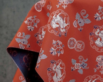 May include: Close-up of a folded fabric with a repeating pattern. The fabric is a vibrant red with a detailed design featuring white and blue floral and animal motifs. The reverse side of the fabric is a deep blue color.