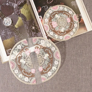 May include: Two round, white, embroidered lace doilies with pink and brown floral designs. The doilies are on a gray fabric background.