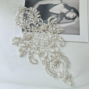 May include: A silver-colored, ornate applique with clear crystal rhinestones. The applique features a floral and leaf design with a mesh backing. The design is embellished with various sizes of sparkling crystals, creating a luxurious look.