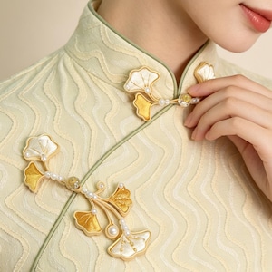 Könnte beinhalten: Ein blassgelbes Kleid mit Stehkragen, strukturiertem Wellenmuster und dekorativen Verschlüssen in Form von Ginkgoblättern. Die Verschlüsse sind mit kleinen weißen Perlen und goldfarbenen Details verziert, was für eine botanische Optik sorgt.