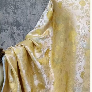 May include: A draped fabric with a floral pattern in yellow and cream hues. The material has a subtle sheen, and the design features detailed botanical elements. The fabric is likely used for home decor or fashion.