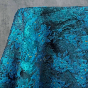 May include: Close-up of a teal fabric with a floral pattern. The design features blue flowers with gold accents against a dark background. The fabric has a textured appearance, suggesting a brocade or similar weave.