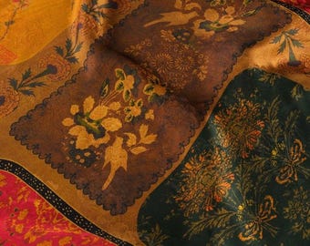 Op de afbeelding: Een stof met een patchwork-ontwerp met verschillende bloemmotieven in tinten goud, bruin, groen, rood en oranje. De stof heeft een textuur, een licht verweerde uitstraling.