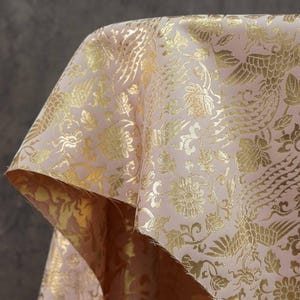 Puede incluir: Primer plano de una tela rosa con un estampado floral y de pájaros dorado. La tela está doblada, revelando un reverso dorado. El diseño presenta representaciones detalladas de pájaros y flores.