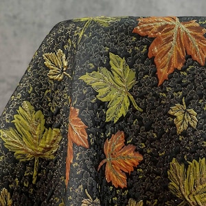 Könnte beinhalten: Schwarzer Stoff mit geprägtem Blumenmuster und gestickten Blättern in Grün, Orange und Gold. Die Blätter haben einen metallischen Glanz und erzeugen ein strukturiertes Design zum Thema Herbst.