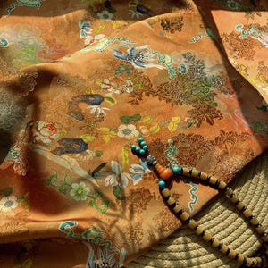 May include: An orange fabric with a floral and bird pattern. The fabric is draped, with a beaded necklace featuring turquoise and orange beads resting on top. The necklace has a wooden bead design.