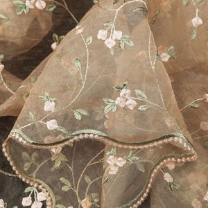 May include: Close-up of a sheer, light brown fabric with delicate floral embroidery. The fabric has a scalloped edge and a subtle sheen.