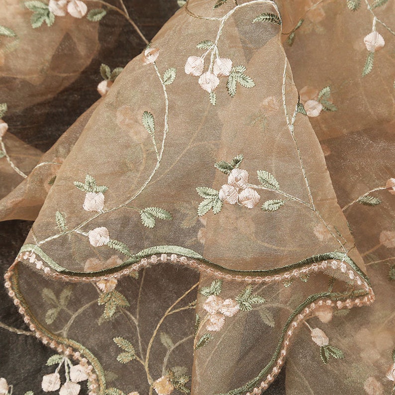 May include: Close-up of a sheer, light brown fabric with delicate floral embroidery. The fabric has a scalloped edge and a subtle sheen.