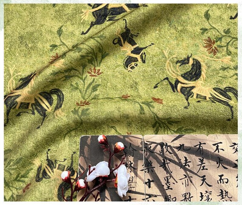 Może przedstawiać: Zielona tkanina z powtarzającym się wzorem czarnych i beżowych koni oraz element&oacute;w kwiatowych. Tkanina ma teksturowany wygląd. Gałązka białych kwiat&oacute;w i kawałek papieru z azjatyckimi znakami znajdują się na pierwszym planie.