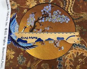 Op de afbeelding: Een close-up van een gouden stof met een blauw en wit geborduurd ontwerp van een kersenbloesemtak. Een houten liniaal met de woorden "Happy Party" ligt op de stof.