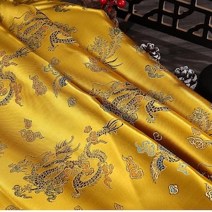 Könnte beinhalten: Goldfarbener Stoff mit einem sich wiederholenden Muster aus Drachen und stilisierten Blumen. Der Stoff hat einen satinartigen Glanz.