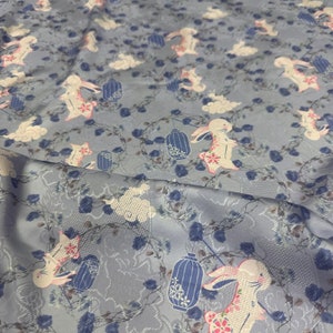 Pode incluir: Um tecido azul claro com um padrão repetido de coelhos brancos segurando lanternas. Os coelhos são cercados por nuvens brancas e flores azuis.