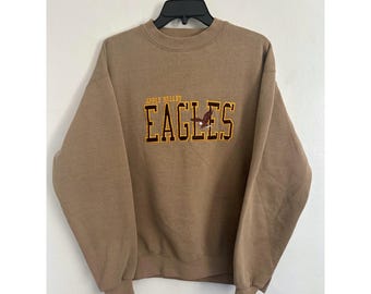 May include: A tan crewneck sweatshirt featuring the embroidered text APPLE VALLEY EAGLES in dark brown with yellow outlines. The letter G contains a small brown eagle graphic. The garment has long sleeves, a ribbed collar, and ribbed cuffs.