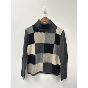 Op de afbeelding: Een grijze coltrui met een patchwork ontwerp. De trui heeft een mix van zwarte, grijze en beige vierkanten. De mouwen en coltrui zijn effen grijs.