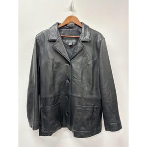 May include: A black leather jacket featuring a notched collar, long sleeves, and a front button closure. The jacket includes two large patch pockets on the lower front section and vertical seam detailing that runs from the waist to the shoulders.