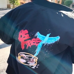 May include: Black bomber jacket with a vibrant design on the back. The design features the words "BE FREE" in red, a blue bird, and a gold-rimmed hourglass. The jacket is a casual outerwear piece.
