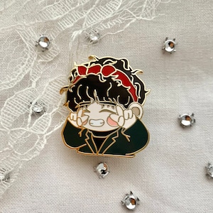 Op de afbeelding: Een gouden emaille pin met een cartoonfiguur met zwart haar, een rode strik en een groen jasje. Het figuur glimlacht en maakt een peace-teken met beide handen.