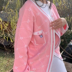 May include: A pink cardigan with a white trim and a heart pattern. The cardigan has a pocket on the left side with a small embroidered cartoon animal.