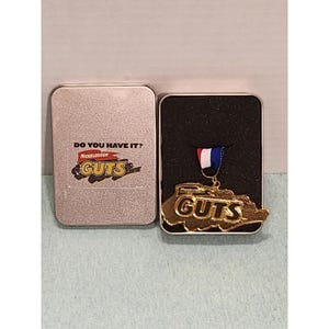 May include: A gold-colored Nickelodeon GUTS medal with a red, white, and blue ribbon, displayed in an open silver tin. The tin lid features the text "DO YOU HAVE IT?" and the GUTS logo.