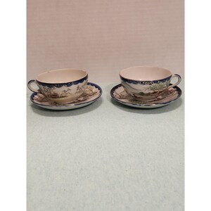 May include: Two matching teacups and saucers. The cups and saucers are white with a blue border and a landscape design. The cups have handles and sit on matching saucers. The design includes trees, mountains, and buildings.