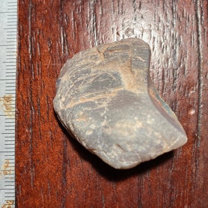 Puede incluir: Primer plano de una roca gris, marrón y roja con una superficie rugosa y texturizada. La roca tiene un diámetro de aproximadamente 5 cm y está sobre una superficie de madera.