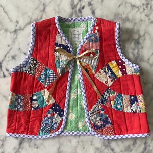 May include: Red quilted vest with a patchwork design and a bow tie closure. The vest has a green and white gingham lining and a blue and white gingham trim. The vest is labeled "House of Quilts".