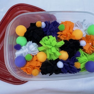 May include: A clear plastic container filled with colourful felt balls and pom-poms. The container is red on the outside and has a clear lid.