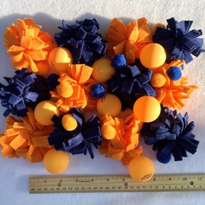 May include: A collection of orange and blue felt toys with orange ping pong balls. The toys are arranged in a cluster on a white surface. A ruler is visible at the bottom of the image.